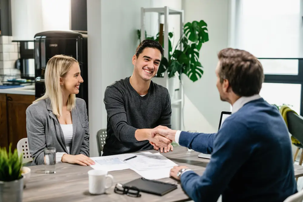 young-happy-couple-making-agreement-with-their-financial-advisor-home-men-are-shaking-hands