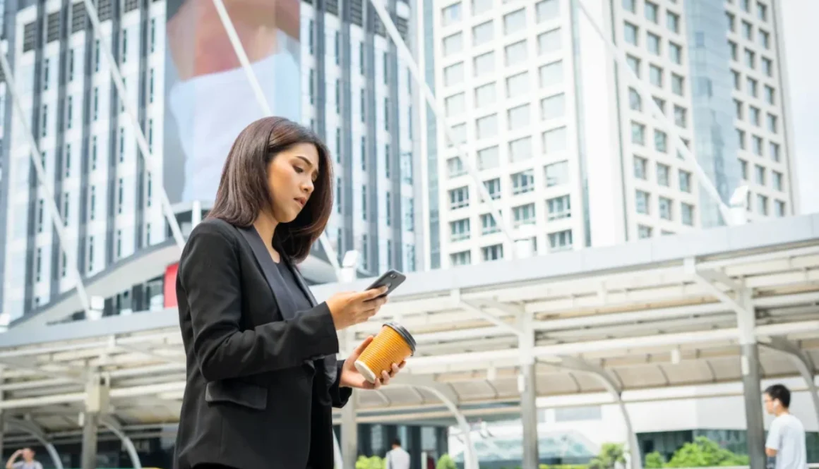 business-woman-using-phone-with-coffee-hand-walking-street-with-office-buildings-background