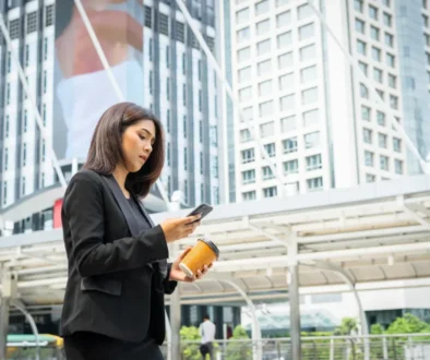 business-woman-using-phone-with-coffee-hand-walking-street-with-office-buildings-background